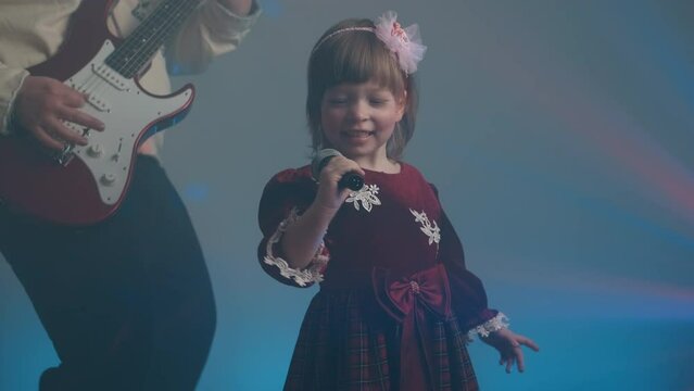 Little girl on stage in vintage dress, she sings into microphone and dances, her father plays an electric guitar. Color music is shining and smoke is billowing. Family time, creativity and hobbies