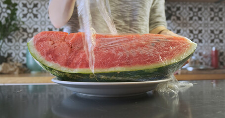 Girl removes a transparent wrapper from a large piece of red juicy watermelon lying on a plate in the kitchen.