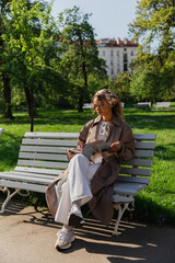 young african american woman in headscarf and trendy outfit sitting on bench and reading magazine.