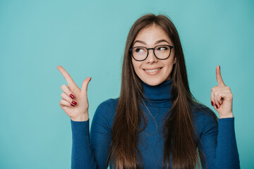 Pretty caucasian woman in blue sweater at studio over turquoise backdrop in glasses, looking up,...