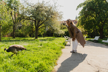 happy african american woman in headscarf and stylish trench coat looking at peacock in park.
