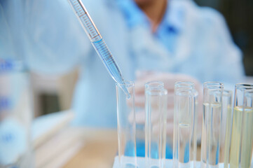 Details: glass dropper with a dripping reagent in a test tube on tripod, over blurred background of a pharmacologist scientist conducting clinical research in the science pharmaceutical laboratory