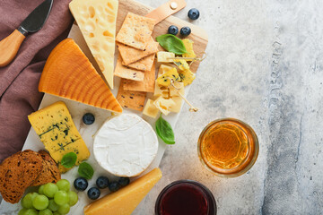 Assortment of cheese, honey, cracker, blueberries, grapes with red and white wine in glasses antipasto server on white marble board on grey background. Flat lay, copy space.