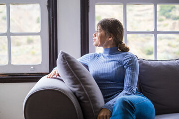 Obraz premium One cute and beautiful young girl looking outdoors at the window from the sofa relaxing and resting. Thoughtful woman pensive and thinking sitting on the couch.