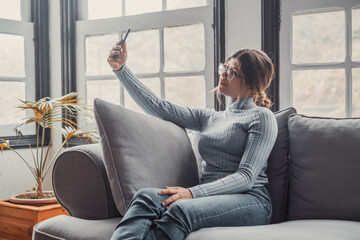 Young beautiful woman sitting on the sofa at home chatting and surfing the net. Female person having fun with smartphone online. Portrait of girl smiling taking a selfie to post it on the social media - obrazy, fototapety, plakaty