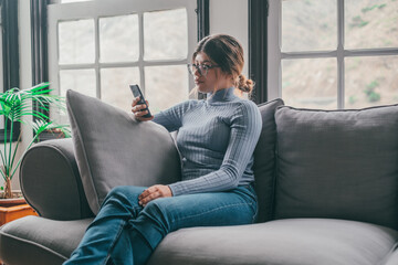 Young beautiful woman sitting on the sofa at home chatting and surfing the net. Female person having fun with smartphone online. Portrait of girl smiling using cellphone revising social media