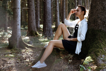 A girl is resting in the woods under a tree. A ribbon in her hair. Trees in the background. The girl is dressed in a light blouse and a black skirt. Sits on the ground, yellow pine needles around
