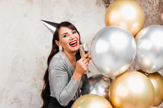 A girl with balloons is laughing against the background of a decorative wall. A beautiful happy woman on the birthday of a New Year's holiday party having fun holding a glass of champagne