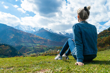 Naklejka premium Close-up of a young woman with a cup of coffee sitting on the grass and admiring the beautiful,incredible view of the mountains on a sunny autumn day.Concept of travel,outdoor.