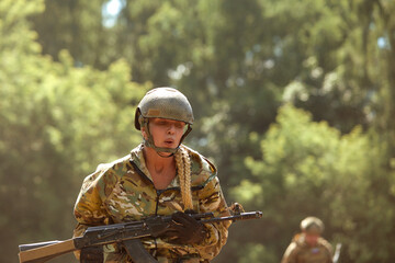 Caucasian Military lady woman in tactical gear posing for photo at summer season. Wearing green camo uniform and assault rifle, in military gear and headset, lady is looking at side