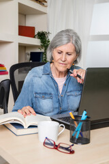Elderly woman looking at a book in front of a computer on a desk