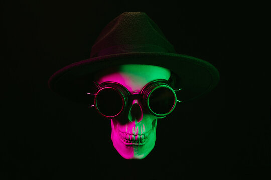 human skull in steampunk glasses and a hat with a pink green light on a dark background