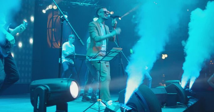 Band performs on stage at musical concert in spotlight and smoke. Vocalist actively moving and gesticulating. Slow motion
