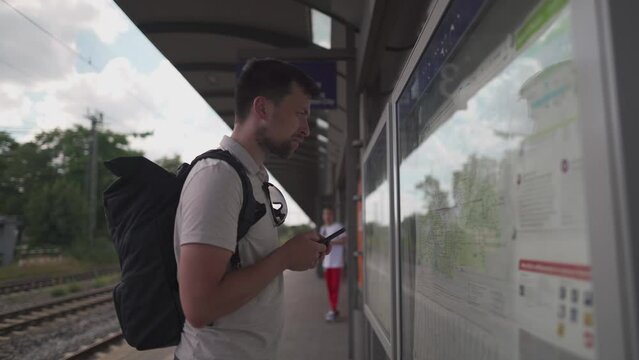 Thoughtful traveler using tourist transport map, looking for best way, transport connection standing at station in Germany, Munich. Man looking for location on mobile phone app and map board at stop 