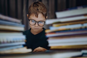 cute caucasian boy wearing glasses reading a book. Cheerful boy peeking from behind piles of books