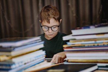 cute caucasian boy wearing glasses covering head with book. Cheerful boy peeking from behind piles of books