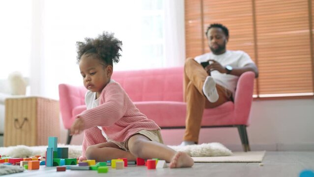 Happy cheerful kid sitting at living room playing with building blocks or wood blacks game with smile. Father sitting on sofa and using phone. Education child toys for preschool and kindergarten. 