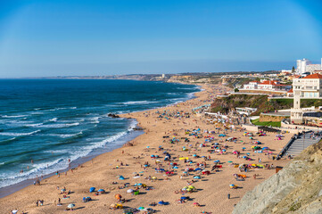 Scenic view of crowded Santa Cruz beach with colorful umbrellas and Atlantic Ocean coastline on a sunny day in Portugal.