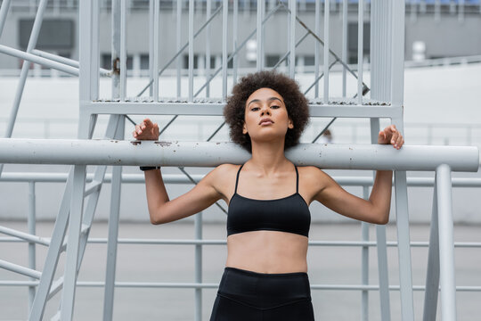 pretty and athletic african american woman in black sports bra looking at camera near fence.