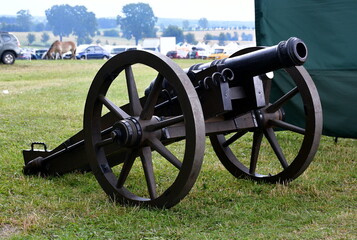 A close up on a replica of a Napoleonian cannon made out of metal and wood standing in the middle of a field or meadow next to a green tent seen during a hike on a Polish countryside