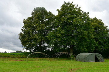 A view of a set of military tents, two bare frames and one covered one, standing next to a dense forest or moor near a vast lawn, meadow, pastureland, or farmland spotted on a cloudy summer day
