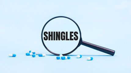 On a blue background, white and blue capsules with pills and a black magnifying glass with the text SHINGLES. Medical concept.