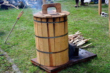 A close up on a wooden barrel with a lid used to store smoking food to add a charred effect to it seen with some smoke going out of it and some firewood nearby seen on a sunny summer day in Poland