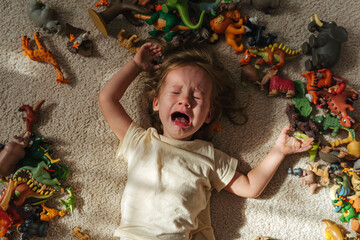 Toddler cries and is naughty lying on the floor among toys in the nursery. Children's tantrums, tears and discontent. Problems of kids upbringing. - obrazy, fototapety, plakaty