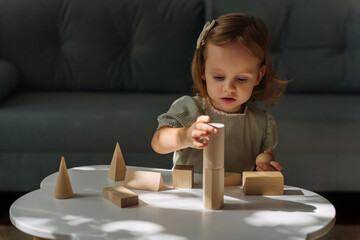 A little girl plays and learns geometric shapes on the table. The kid builds a tower from wooden ...