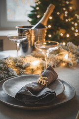 Christmas festive table setting with garland, champagne, stylish gray plate with napkin ring. Family holiday dinner home party. Vertical format. Close up.