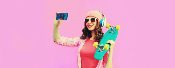 Summer colorful portrait of stylish modern young woman taking a selfie by smartphone with skateboard and headphones on pink background