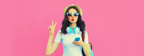Portrait of stylish young woman in headphones listening to music with smartphone and blowing her lips on pink background