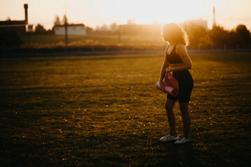 athletic curly woman laying a gymnastic mat before training outdoors at sunset.