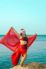 A woman in a costume for oriental dances in a Santa hat is dancing with a headscarf on a rock against the background of the sea.