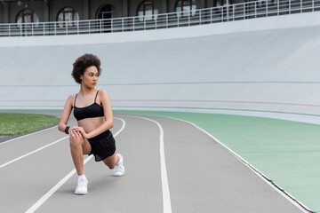 Fototapeta premium Young african american sportswoman doing lunges on running track of stadium.