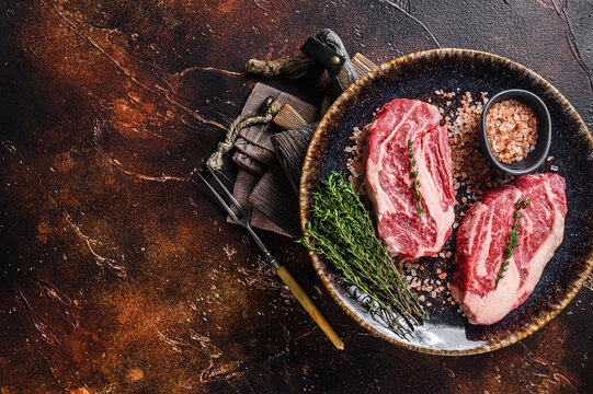 Chuck eye Roll beef steaks, raw meat  on a plate with thyme and herbs. Dark background. Top view. Copy space