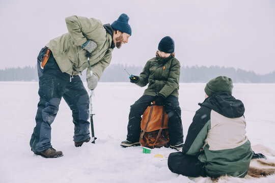 Cheerful man using ice auger while talking with backpack at frozen lake