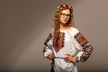 A beautiful and young model stands on a gray background. The model is turned sideways to the camera and is wearing national clothes. On the head is a wreath of flowers and glasses. naturally beautiful