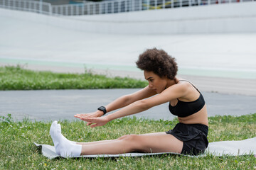 Fototapeta na wymiar Young african american sportswoman with smart watch stretching on fitness mat on stadium.