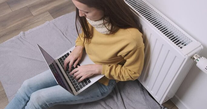 Woman works on laptop near battery. Woman freelancer in scarf warms herself near radiator. Sitting in room try warming indoor. Cold house, poor central heating, low temperature in apartment concept