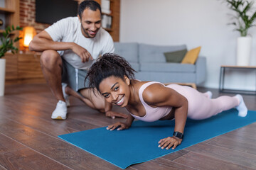 Cheerful millennial african american man in sportswear helping to wife doing push ups in living room interior