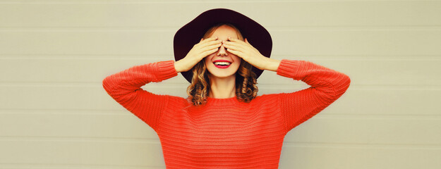 Autumn portrait of cheerful happy laughing woman covering her face with her hands wearing hat, red knitted sweater on gray background