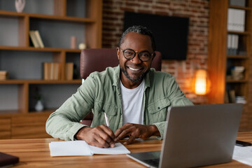 Smiling middle aged african american guy in glasses works on laptop at table, makes note, at table...