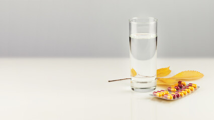 Health care autumn banner with pills, full glass of pure water and autumn leaves on white table with copy space. Concept of flu season and no virus. Vitamins and nutrients in capsules for good health