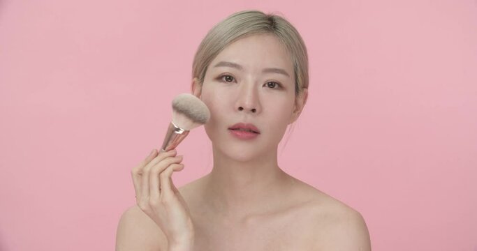 Beauty portrait of young pretty Asian woman with blond hair makes blush on the cheek and using makeup brush runs over her face, looking and smiling at camera on isolate pink background. 
