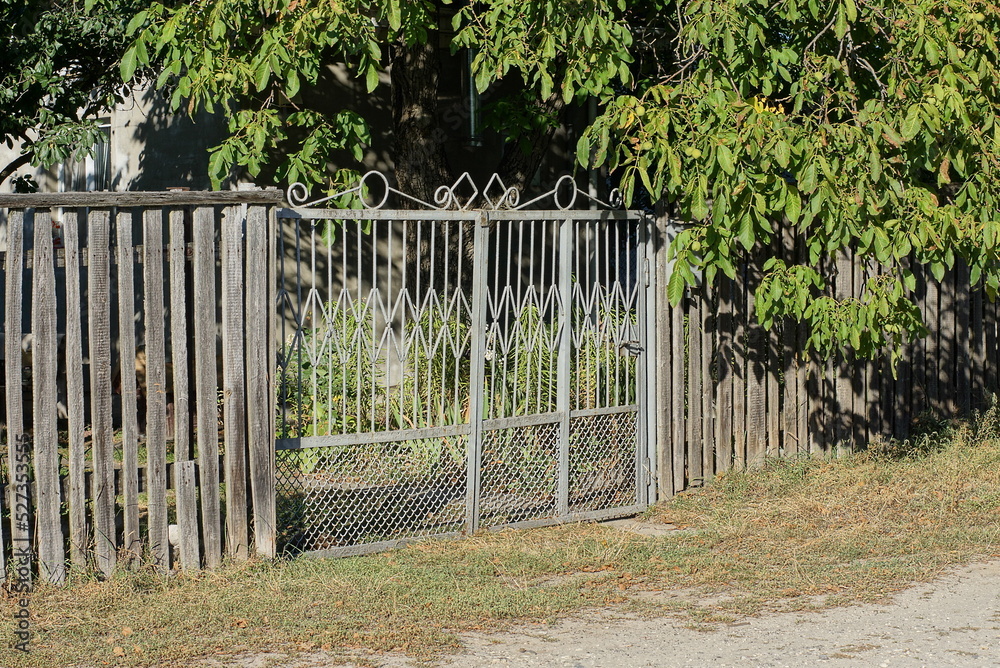 Sticker one closed gray metal gate on a wooden fence wall overgrown with green vegetation on a rural street