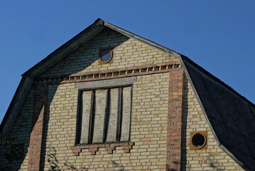 one attic of an old brown abandoned brick private house with a window boarded up with plywood under a gray slate roof against a blue sky