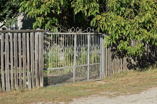 one closed gray metal gate on a wooden fence wall overgrown with green vegetation on a rural street