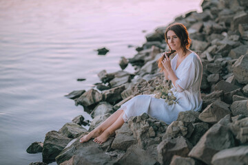 Sensual girl in white long vintage dress makes a braid near the river on stones. Harmony and rest with nature. Ukrainian tradition celebration of Ivana Kupala. Relax, travel, quiet place