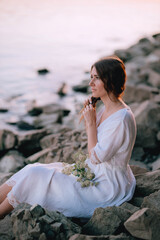 Fototapeta na wymiar Sensual girl in white long vintage dress makes a braid near the river on stones. Harmony and rest with nature. Ukrainian tradition celebration of Ivana Kupala. Relax, travel, quiet place